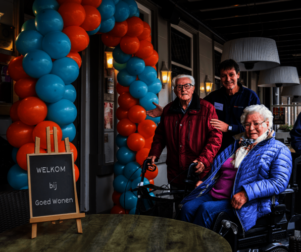 De ingang met ballonnenboog. Waar buurtcoach Mark twee trouwe huurders welkom heet. Vrouw in een rolstoel en man staand met rollator.