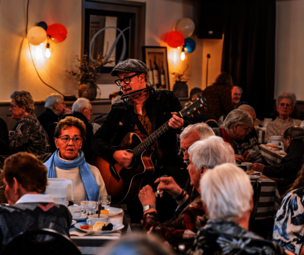 Troubadour Leo die bij een tafel met huurders staat te zingen en gitaar speelt.