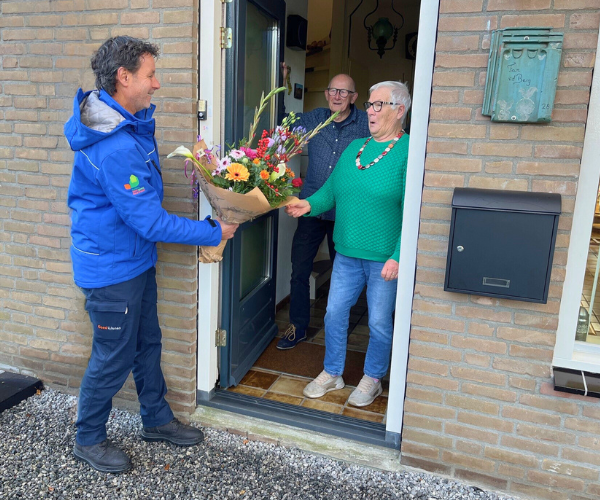 De heer en mevrouw Van den Berg in de deuropening van hun woning. Mevrouw neemt een bos bloemen aan van buurtcoach Mark.