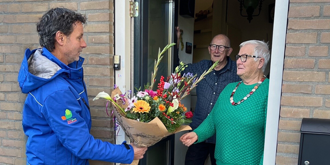De heer en mevrouw Van den Berg in de deuropening van hun woning. Mevrouw neemt een bos bloemen aan van buurtcoach Mark.
