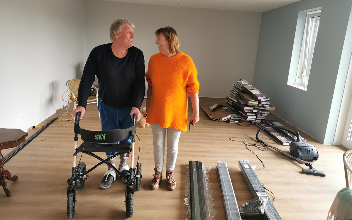 Ada en Peel kijken elkaar lachend aan in hun appartement, met om hen heen klusspullen. Peel staat met een rollator.