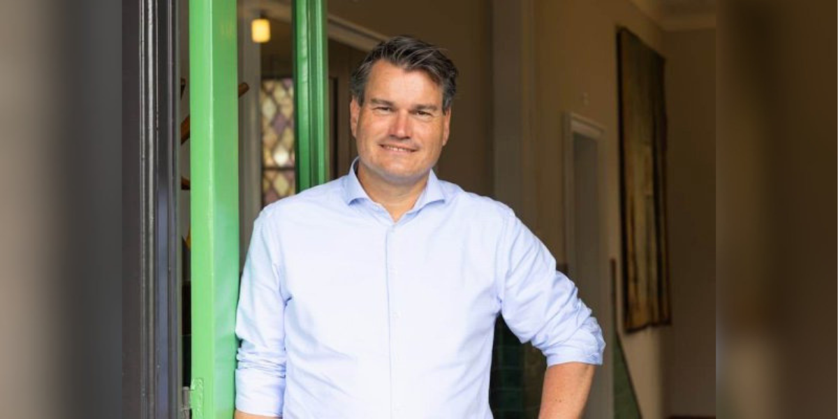 Foto van Dries Ewalds vanaf middel omhoog. Met lichtblauwe blouse en glimlach. In een deuroperning, leunend tegen groene deurstijl. 