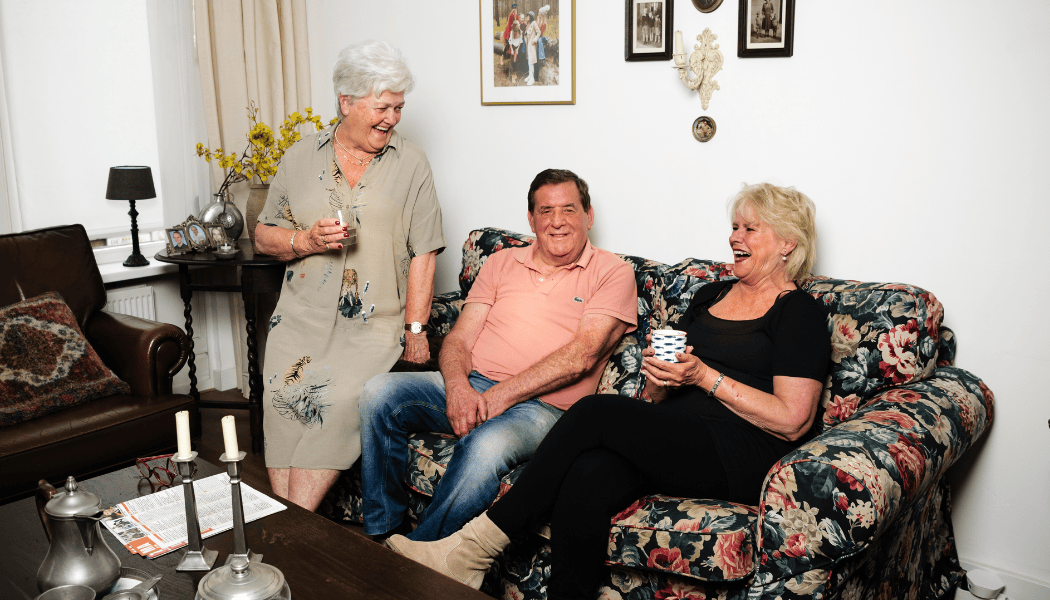 Foto van een woonkamer met een senioren stel op de bank en een vijftiger. Lachend.