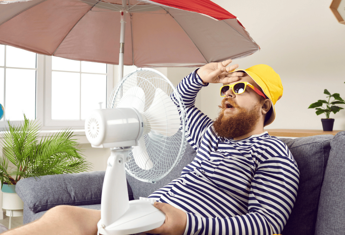 Man in een blauw wit gestreept shirt en geel hoedje zwetend op de bank met een rode parasol en ventilator