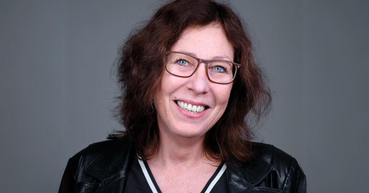 Joke Abbring met een grote glimlach en bril. Foto vanaf borsthoogte omhoog.
