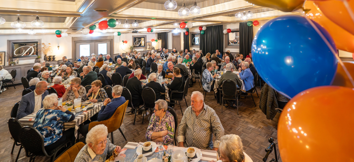 Overzichtsfoto van jubileumlunch met tientallen oudere huurders aan tafeltjes in een grote zaal. 
