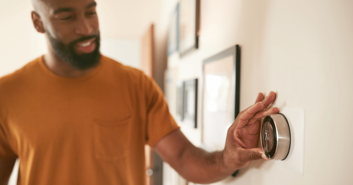 Een man met een donker huidskleur een baard en snor en oranje shirt draait aan de thermostaat in een woning. Met tekst 'Noodfonds Energie' op de foto.
