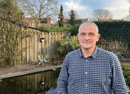 Tom van Woerden vanaf borsthoogte staande in een achtertuin voor een vijver, schutting en groen.