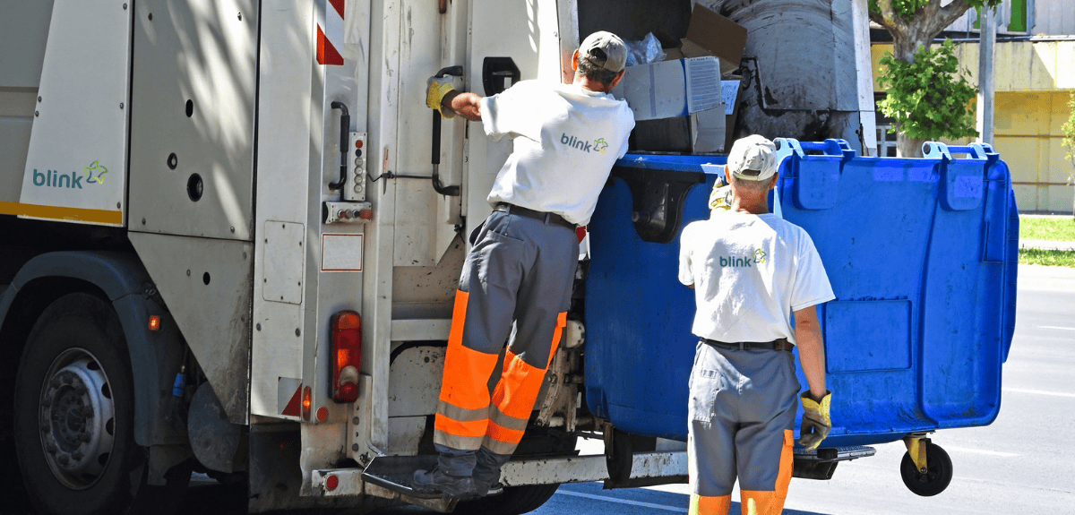 Twee vuilnismannen zijn bezig een blauwe container in een vuilniswagen te ledigen.