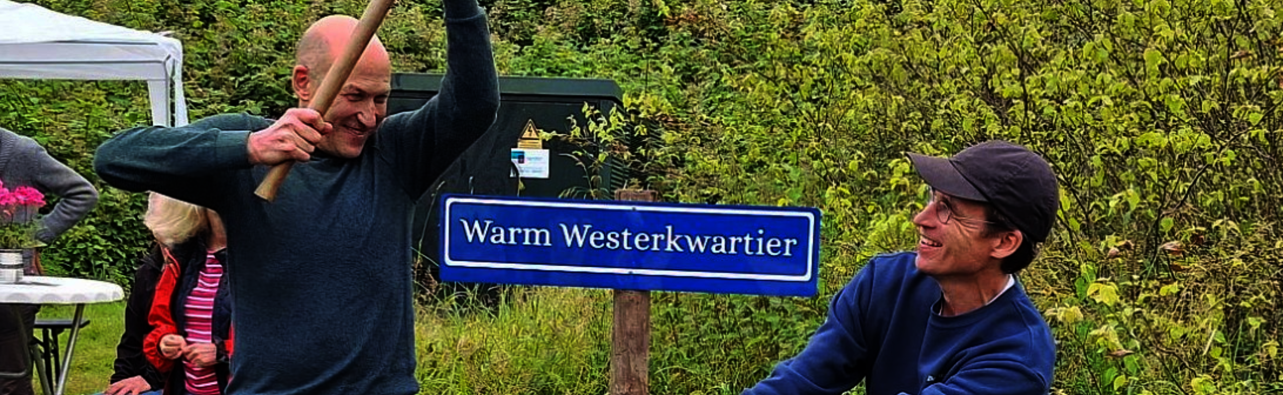 Een man houd een hamer in de lucht waarmee hij een bordje in de grond slaat waarop staat""Warm westerkwartier" een andere man houdt het plaatje onder het bordje vast. 