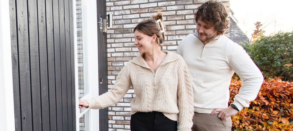 Twee personen staan bij de voordeur van een woning; één persoon opent de deur terwijl de ander naast staat, met een gevel van baksteen en herfstkleurige struiken op de achtergrond.