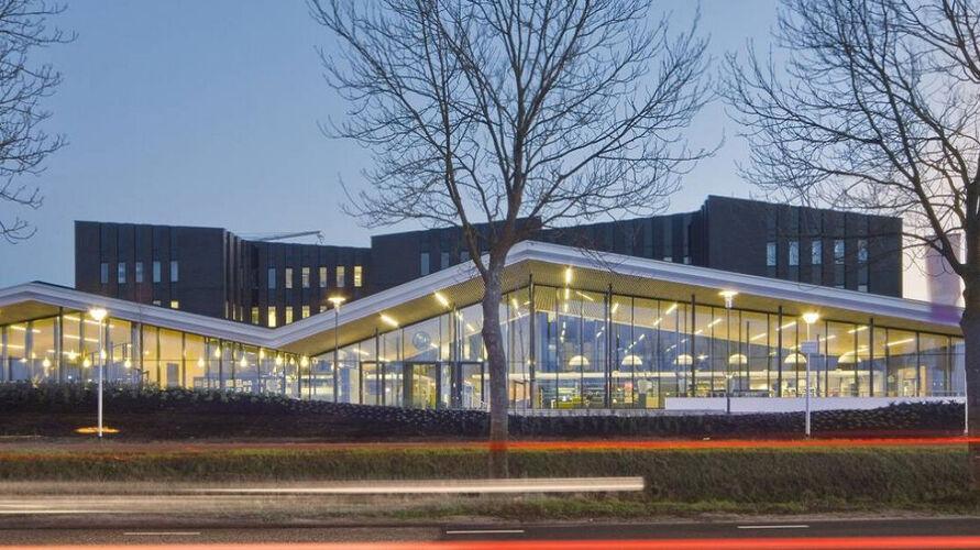 Gemeentehuis Rucphen. Modern gebouw met grote glazen ramen en een hoekig dak, verlicht van binnen. Op de voorgrond staan kale bomen en lichtsporen van passerende voertuigen zijn zichtbaar tegen een blauwe schemerlucht.