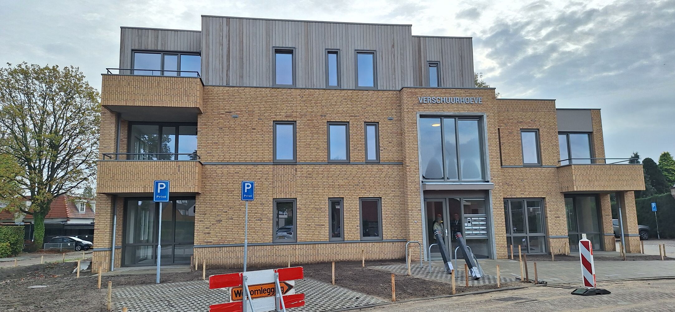 Een modern gebouw met bakstenen gevel, grote ramen en meerdere verdiepingen. Aan de voorkant hangt een bord met 'Verschuurhoeve'. Er zijn gehandicaptenparkeerplaatsen gemarkeerd met blauwe borden.
