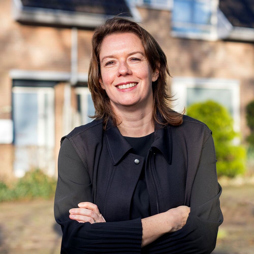 Danielle Gerrits, de nieuwe directeur bestuurder van Thuisvester met zwarte kleding, met gekruiste armen buiten voor een woning met bakstenen gevel en ramen