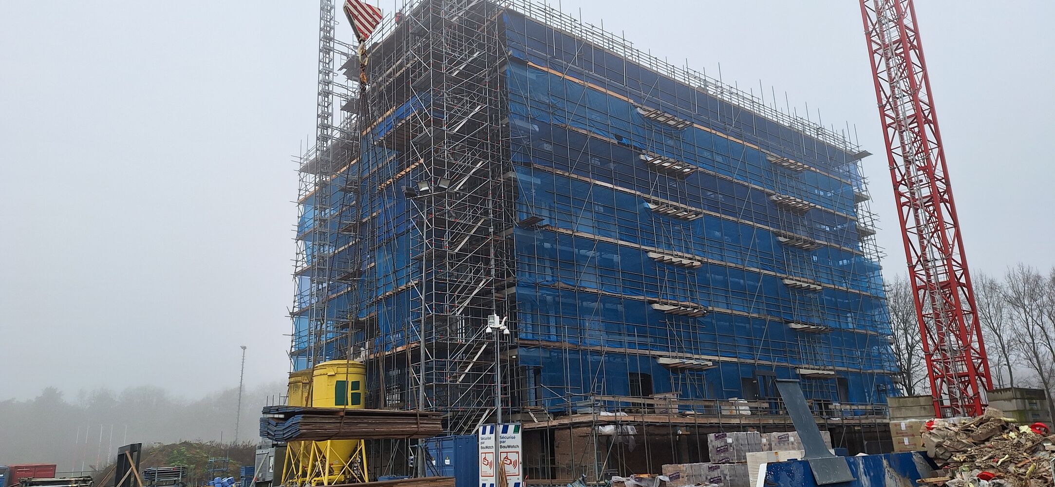 Gebouw in aanbouw bij Parck Warande in Oosterhout, omgeven door steigers en blauwe netten, met een rode bouwkraan en een Amerikaanse vlag bovenop.