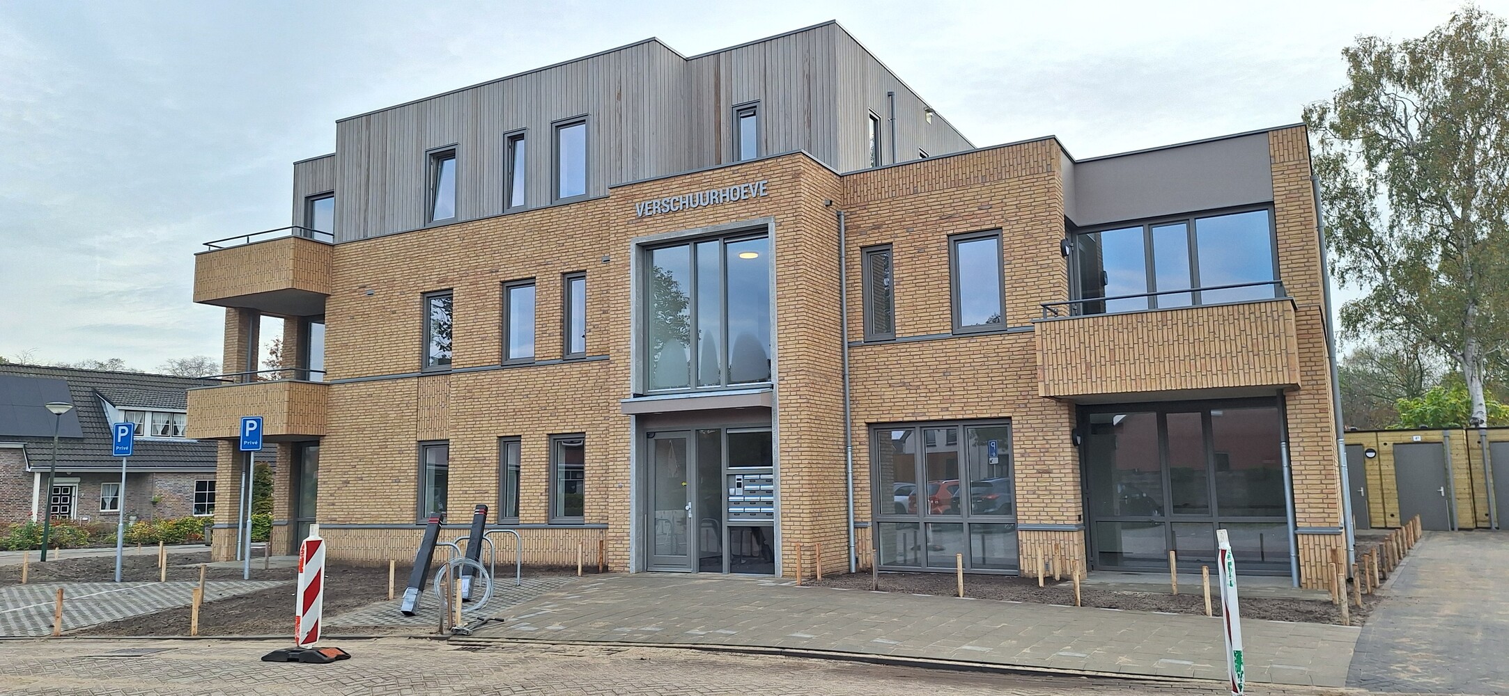 Vooraanzicht van een modern gebouw met bakstenen en houten gevel, voorzien van grote ramen en een bord met de tekst “Verschuurhoeve"