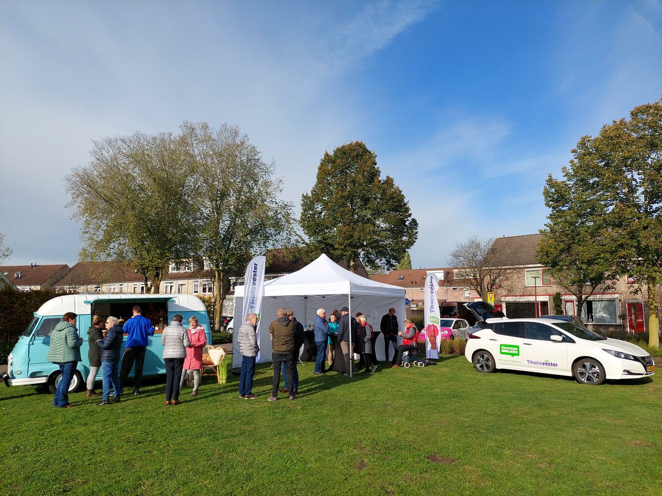 Groep mensen bij witte tent en voertuigen op grasveld