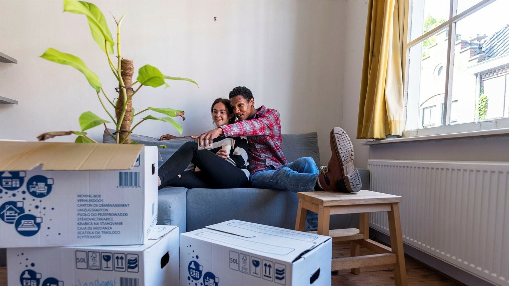 Twee personen zitten op een grijze bank in een woonkamer met verhuisdozen op de voorgrond en een grote raam aan de rechterzijde