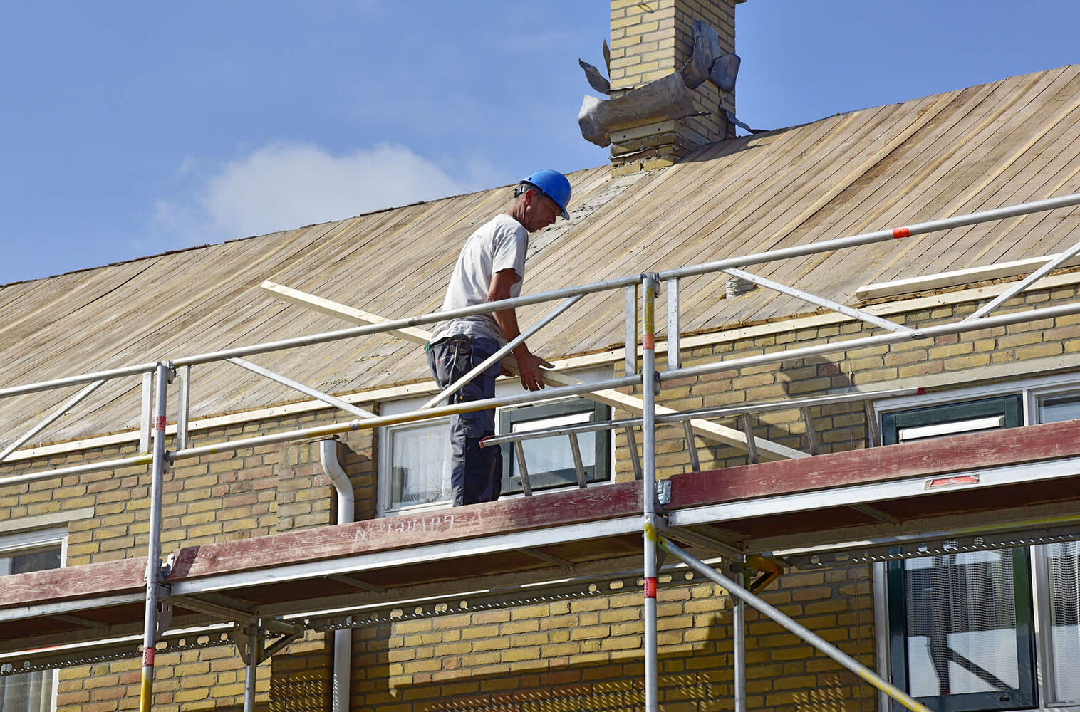 Bouwvakker op steiger werkt aan dakrenovatie van een huis met gele bakstenen gevel en schoorsteen.