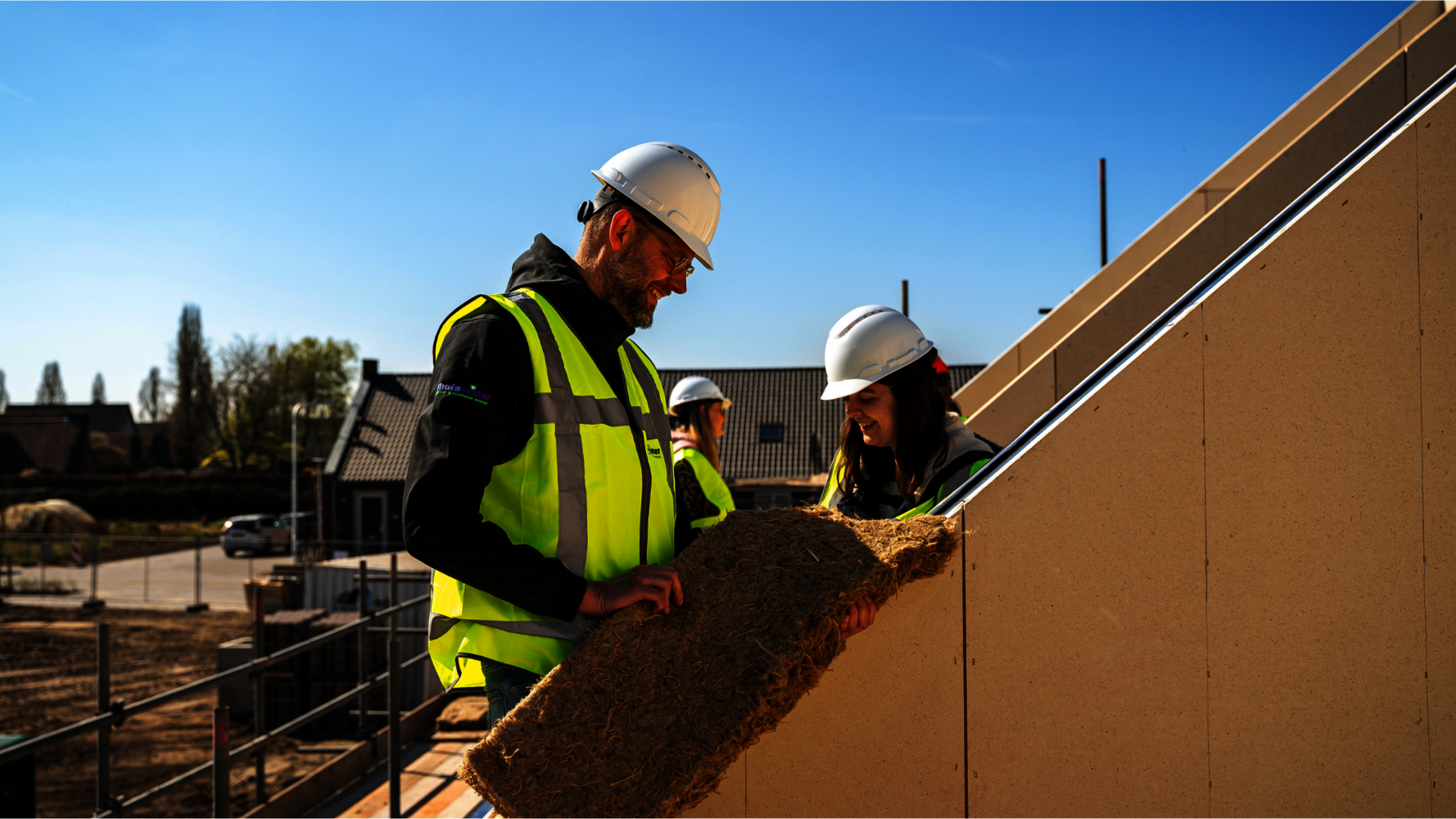 Twee personen met witte bouwhelmen en veiligheidsvesten plaatsen isolatiemateriaal tegen een dakconstructie op een bouwplaats.