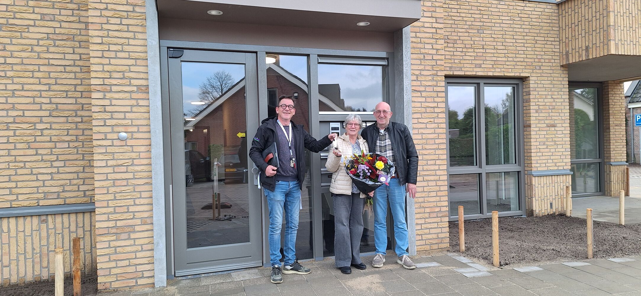 Drie mensen staan buiten voor een modern gebouw met lichtkleurige bakstenen en grote ramen. Eén persoon houdt een kleurrijk boeket bloemen vast, wat wijst op een feestelijke gelegenheid. De sfeer is informeel en vriendelijk.