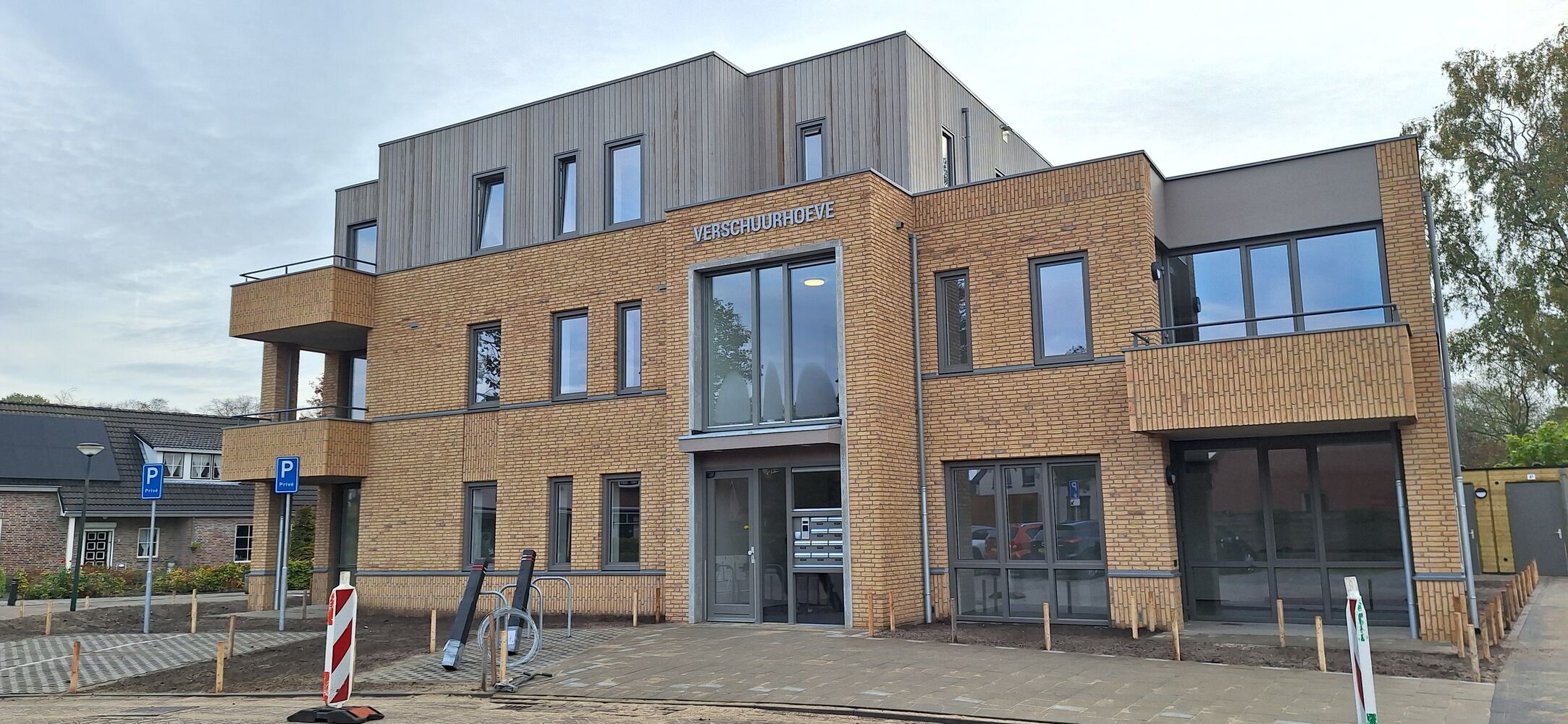Een modern gebouw met bakstenen gevel, grote ramen, meerdere verdiepingen en balkons. Aan de voorkant hangt een bord met de tekst 'Verschuurhoeve'. Voor het gebouw zijn twee gehandicaptenparkeerplaatsen gemarkeerd.