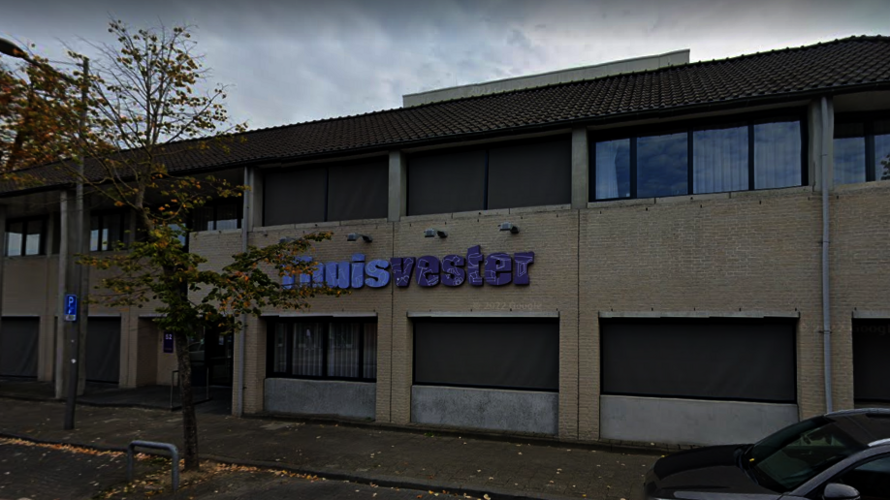 Twee verdiepingen tellend gebouw met beige bakstenen gevel en donker pannendak, met het paarse logo "Thuisvester" op de voorgevel. Er staan auto's geparkeerd langs de straat en bomen sieren het trottoir.