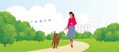 Persoon met rode hoodie wandelt met een hond aan de lijn over een pad in het park, met bomen en gebouwen op de achtergrond.