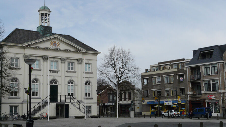 Gemeentehuis Zundert, een wit historisch gebouw links, voorzien van een torentje en trap naar de ingang, omringd door kleinere winkels en appartementen.
