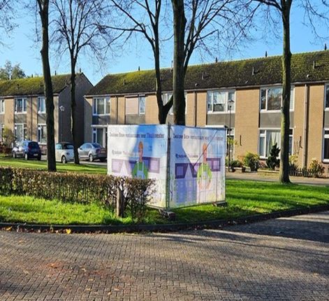 Straat met rijtjeswoningen en een groot informatiebord over een energieproject, geplaatst op een grasstrook tussen bomen